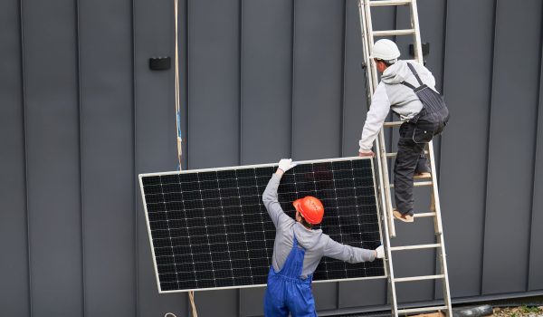 Twee werkers die een zonnepaneel vasthouden, een staat op de grond en de ander op een ladder